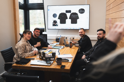 Meeting room with people at Vanquish Fitness around a wooden table, laptops and notebooks, and a screen showing black t-shirt designs