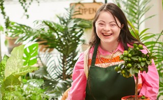 a white woman with down syndrome, wearing a pink shirt and green apron, smiling as she works with plants in a plant shop
